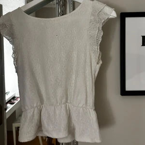 Vit spetsblus - Säljer en söt vit spetsblus med volangdetaljer. Blusen är ärmlös och perfekt för sommarens alla tillfällen. Den har en feminin och romantisk stil som passar både till vardags och fest. Superfin att matcha med jeans eller kjol!