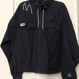 Svart vindjacka från Nike jordan - Säljer en svart vindjacka från Nike jordan med dragkedja och huva som man kan rulla och gömma i jackan. Jackan har en cool design med vita detaljer och en liten logga på bröstet. Perfekt för blåsiga dagar och sportiga aktiviteter. Passar både till vardags och träning!