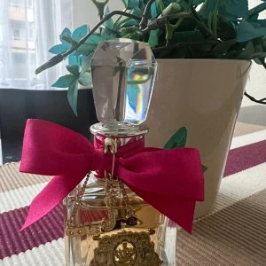 Parfym från Juicy Couture - Säljer en lyxig parfym från Juicy Couture. Flaskan är elegant med en stor, glittrande kristallkork och en vacker rosa rosett runt halsen. Den har en gyllene etikett med detaljerad design. Perfekt för den som älskar en glamorös stil!