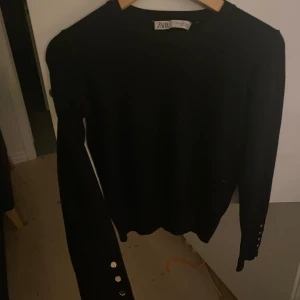 Svart tröja med knappar från Zara - Säljer en stilren svart tröja från Zara. Den har långa ärmar med snygga guldfärgade knappar längs ärmsluten. Perfekt för både vardag och fest! 🖤