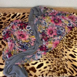 Blommig halsduk/slöja i grått och rosa - Säljer en superfin blommig halsduk med grå bas och färgglada blommor i rosa, blått och gult. Perfekt för att liva upp din outfit under höst och vår. Materialet är lätt och luftigt, vilket gör den skön att bära. Passar både till vardags och fest! 🌸