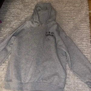 Grå hoodie från Gina Tricot - Mysig grå hoodie från Gina Tricot med texten 'JE M'EN AMOUR' och en stjärna tryckt på framsidan. Perfekt för en avslappnad stil och passar bra till både jeans och leggings. Den har en klassisk passform med långa ärmar och en bekväm huva.