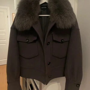 Grå jacka från Meotine - Säljer en snygg grå jacka från Meotine med en lyxig pälskrage i blå räv. Jackan har en blandning av ull och polyester, vilket gör den både varm och hållbar. Den har knappar framtill och praktiska fickor. Perfekt för höst och vinter!