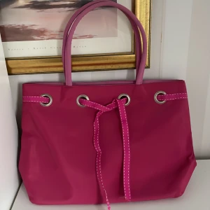 Rosa axelväska med knytband ny - Säljer en superfin rosa axelväska i syntetmaterial. Den har ett snyggt knytband framtill och silverfärgade öljetter. Perfekt för vår och sommar! Väskan har en praktisk dragkedja på baksidan för extra förvaring.