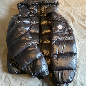 Svart pufferjacka från Moncler - Säljer en snygg svart pufferjacka från Moncler. Den är perfekt för kalla dagar med sitt blanka material och varma fyllning. Jackan har en dragkedja framtill och en cool logga på bröstet. Den är i bra skick och redo för nya äventyr! 