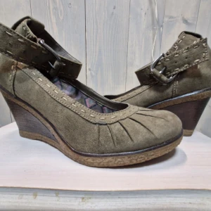 Olivgröna mocka kilklackar - Snygga olivgröna kilklackar i mocka med dekorativa sömmar och justerbart spänne. Perfekta för höst och vår, dessa skor har en stabil kilklack och en bekväm passform. Insidan har ett mönstrat foder som ger extra stilpoäng. Perfekta för både vardag och fest!