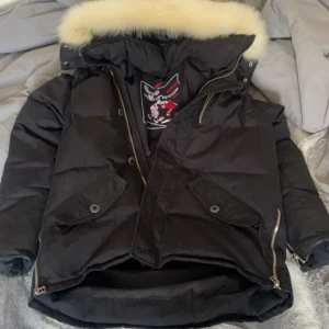 Svart dunjacka från Moose Knuckles - Säljer en svart dunjacka från Moose Knuckles, perfekt för kalla dagar. Jackan har en broderad logga och dragkedja samt knappar framtill. Den är inte i bra skick reva i ärmen och i fickan . Passar perfekt för vintern och håller dig varm och stilren.