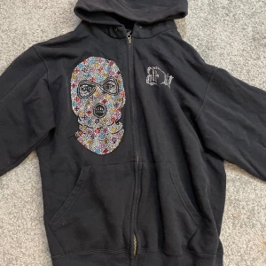 Svart hoodie med strassdetaljer - Cool svart hoodie med en stor strassdesign av en maskerad figur på framsidan och texten 'BANK ROBBER' i strass på baksidan. Köpt för 1100kr