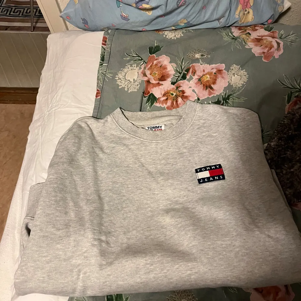 Säljer en stilren grå sweatshirt från Tommy Jeans i storlek M. Tröjan har en klassisk design med ribbade muddar och en liten logga på bröstet. Perfekt för en avslappnad look eller som ett lager under jackan. Passar både höst och vår!. Tröjor & Koftor.