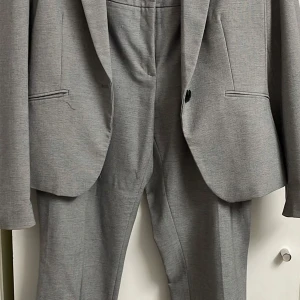 Grå kostym i två delar - Grå kostym från H&M i två delar. Kavajen har en klassisk design med en knapp och byxorna har en rak passform. Perfekt för både kontoret och mer formella tillfällen. Materialet är mjukt och bekvämt, vilket gör den idealisk för långa dagar.