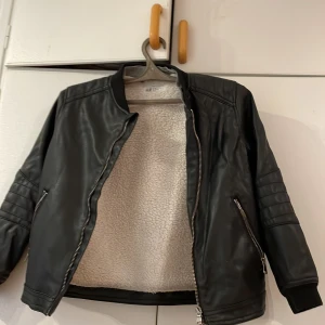 Svart skinnjacka från H&M - Säljer en snygg svart skinnjacka från H&M i herrstorlek M. Jackan har en mjuk och varm foder i vitt teddy-material, perfekt för höst och vår. Den har dragkedja framtill och två fickor med dragkedjor. Ärmarna är långärmade med quiltade detaljer. Perfekt för en cool och stilren look!