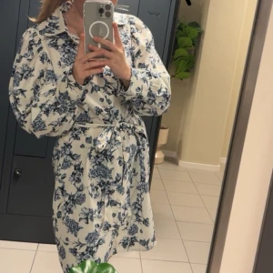 Blommig klänning med knytband - Säljer en blommig klänning med knytband i midjan å puffärm. Har ett vackert blått blommönster på vit bakgrund. Perfekt för en avslappnad och chic look.