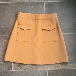 Beige/orange kjol med fickor - Orange kjol med två framfickor med knappar och en dragkedja bak. Längd 43 cm & midja 36 cm.