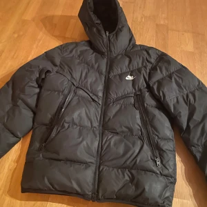 Svart pufferjacka från Nike - Säljer en svart pufferjacka från Nike med dragkedja och huva. Jackan har en sportig look med två praktiska fickor framtill. Perfekt för kyliga dagar.
