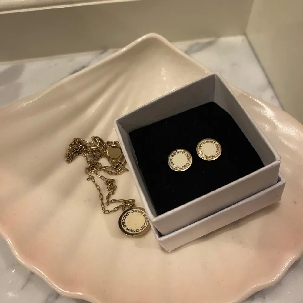 Elegant set med ett guldhalsband och matchande örhängen från Marc Jacobs. Halsbandet har en rund hängande berlock med märkets namn ingraverat. Örhängena är runda och matchar perfekt till!. Asusteet.