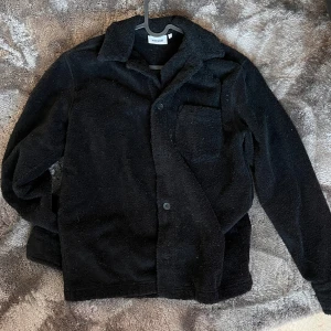 Svart fleecejacka från Weekday -  svart fleecejacka från Weekday med knappar framtill och en bröstficka. Perfekt för kyliga dagar när du vill hålla dig varm och bekväm. Jackan har en klassisk krage och långa ärmar. 