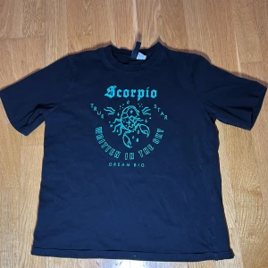 Svart t-shirt med skorpiontryck - Cool svart t-shirt från Divided med ett turkost skorpiontryck och texten 'Scorpio' på framsidan. Perfekt för stjärnteckenentusiaster som vill sticka ut. Kortärmad och i bekväm bomull.