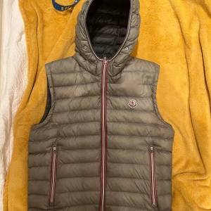 Grå magila cardigan väst  - Snygg grå dunväst från Moncler med huva och dragkedja. Västen har två fickor med dragkedjor och Moncler-logga på bröstet. Perfekt för kyligare dagar. Självklart äkta igensydd dragkedja går inte att dra hela vägen ner