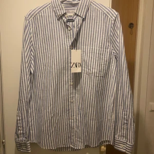 Randig skjorta från Zara - Snygg blå- och vitrandig skjorta från Zara med klassisk krage och bröstficka. Perfekt för en stilren look. Skjortan har långa ärmar och knappar framtill. Storlek S, men väldigt stor i storleken så passar även M. Helt oanvänd och tagen ligger kvar 