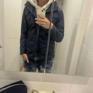 Mörkblå jacka J Lindberg  - Snygg mörkblå jacka från J Lindberg med dragkedja och knappar. Jackan har en hög krage och flera praktiska fickor. Perfekt för höst och vår. Även luva får man 