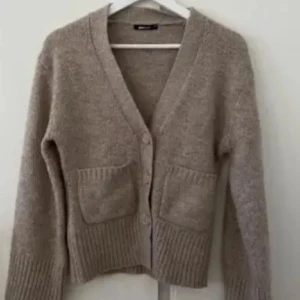 Beige stickad kofta med knappar - Mysig beige stickad kofta med knappar framtill och två fickor. Perfekt för kyliga dagar med sin ribbade nederkant och ärmslut. En klassisk och stilren design som passar till det mesta.