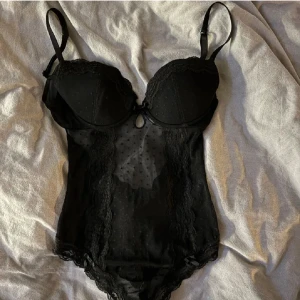 Svart spetsbody - Säljer min svarta body med spetsdetaljer och justerbara axelband. Den har en transparent prickig design framtill och en öppning vid bak. Perfekt nu till nyår! 🎊
