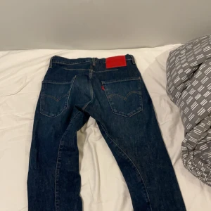 Blå jeansbyxor med röd etikett - Säljer ett par blå levi’s jeans 512. Byxorna har en normal passform och är utrustade med dragkedja och knappar. Perfekta för en avslappnad stil. 