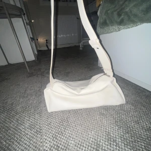 Vit axelväska med justerbar rem - Snygg vit axelväska i syntetmaterial med en justerbar rem och dragkedja. Perfekt för att bära dina nödvändigheter med stil.