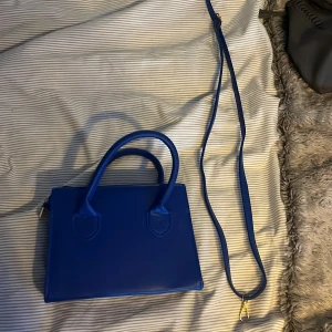 Blå axelväska med dragkedja - Snygg blå axelväska i syntetmaterial med både handtag och en lång axelrem. Väskan har en praktisk dragkedja upptill för säker förvaring. Perfekt för att bära med sig det viktigaste med stil.