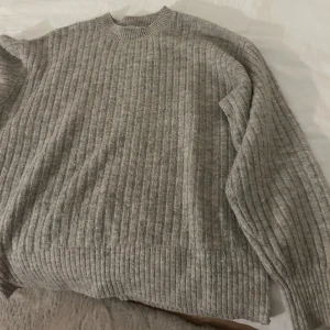 Grå ribbad stickad tröja - Mysig grå stickad tröja med ribbat mönster. Perfekt för kyliga dagar med sin klassiska design och bekväma passform.