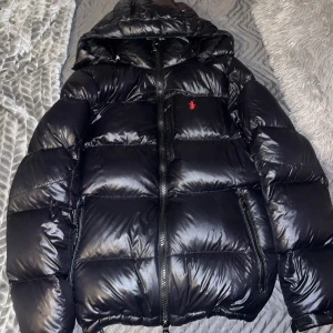 Marinblå dunjacka från Polo Ralph Lauren - Snygg svart dunjacka från Polo Ralph Lauren med huva och dragkedja. Jackan har en klassisk pufferdesign och är perfekt för kyliga dagar. Den har en liten röd logotyp på bröstet som ger en stilren touch.