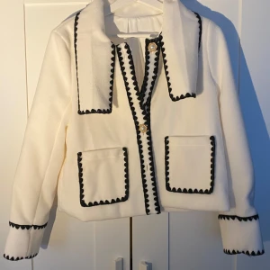 Vit kappa med svarta detaljer - Elegant vit kappa med svarta kantdetaljer och guldknappar. Den har två framfickor och en unik krage. Perfekt för en stilren look.