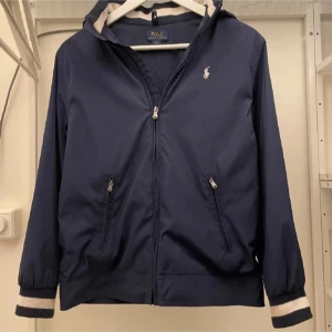 Blå vindjacka från Polo Ralph Lauren - Snygg blå vindjacka från Polo Ralph Lauren med dragkedja och huva. Jackan har långa ärmar med ribbade muddar och vita detaljer. Perfekt för blåsiga dagar. Priset är inte hugget i sten, vid snabb affär kan det sänkas.
