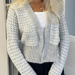 Grå stickad kofta - Mysig grå stickad kofta med ett unikt mönster. Koftan har en öppen design och långa ärmar, perfekt för kyliga dagar. Den ribbade kanten ger en snygg detalj och extra komfort.