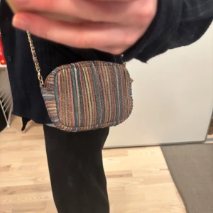 Glittrig randig axelväska Becksöndergaard - Snygg axelväska med glittriga ränder i flera färger. Väskan har en guldfärgad kedja som axelrem och stängs med dragkedja. Perfekt för att lägga till lite färg och glitter till din outfit. Från becksöndergaard. Aldrig använd. 