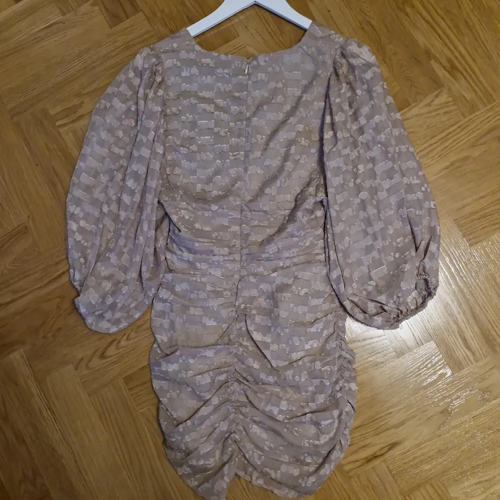 Säljer en helt oanvänd beige klänning från NA-KD med ett unikt mönster och voluminösa trekvartsärmar. Klänningen har en v-ringad design och rynkade detaljer som ger en smickrande passform. Perfekt för speciella tillfällen! Obs liten i storlek. Mekot.