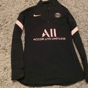 Svart långärmad tröja från Nike med PSG-logga - Säljer en svart långärmad tröja från Nike med PSG-logga och texten 'Accor Live Limitless' på framsidan. Tröjan har rosa detaljer och en halv dragkedja vid halsen. Perfekt för fotbollsfans! ❤️