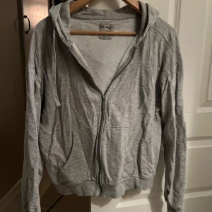 Grå hoodie med dragkedja - Säljer en bekväm grå hoodie med dragkedja och huva. Perfekt för en avslappnad stil. Den har långa ärmar och ribbade muddar vid ärmslut och nederkant.