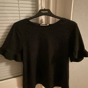 Svart glittrig topp från NA-KD - Snygg svart glittrig topp från NA-KD med korta ärmar och volangdetaljer. Perfekt för en kväll ute eller en festlig tillställning. ✨