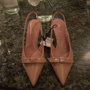 Baby rosa klackar  med rosett från Zara - Eleganta rosa klackar från Zara med spetsig tå och en söt rosett framtill. Skorna har en smal klack och en öppen häl med band. Helt nya och helt oanvända! De är helt slut på lagret! Är också öppen för att byta till en storlek 38! 