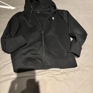 Zip Ralph Lauren - Säljer en stilren svart hoodie från Ralph Lauren med dragkedja och klassisk logga på bröstet. Tröjan har en bekväm passform och praktiska fickor framtill. Perfekt för en avslappnad look.