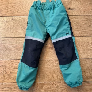 Turkosa och svarta byxor från FIX - Säljer ett par turkosa och svarta byxor från FIX med elastisk midja och muddar vid bensluten. Byxorna har reflexdetaljer och är perfekta för utomhusaktiviteter.
