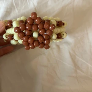 Brunt och vit/beige Kandi armband  - Unikt handgjort armband med bruna och beige pärlor i ett vackert mönster. Perfekt för att ge en bohemisk touch till din stil. Armbandet är elastiskt och passar de flesta handleder.