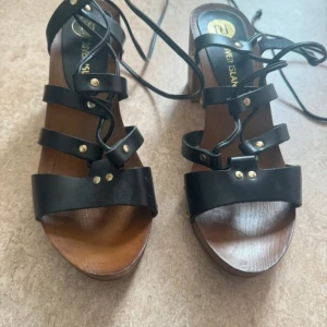 Svarta sandaler med klack och snörning - Snygga svarta sandaler från River Island med klack och snörning. Sandalerna har dekorativa nitar och en robust träliknande sula. Perfekta för en stilren look.
