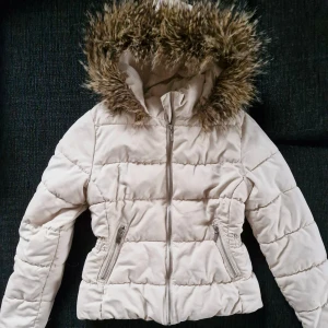 Beige pufferjacka med fuskpäls - Säljer en snygg beige pufferjacka med fuskpäls på huvan. Jackan har dragkedja framtill och två praktiska fickor med dragkedjor. Perfekt för kyliga dagar!
