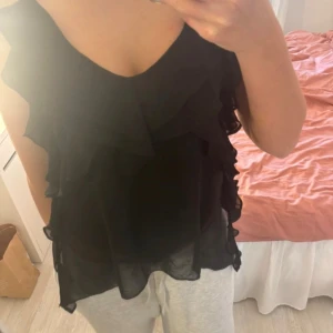 Svart volanglinne - Säljer ett svart linne med volanger. Perfekt för en stilren look. Linnet har en luftig passform och ärmlös design.