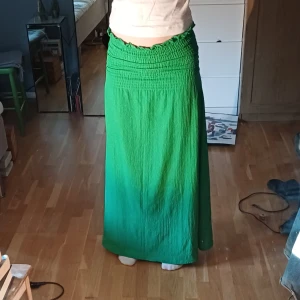 Grön långkjol/klänning - Snygg grön långkjol med elastisk midja som ger en bekväm passform. Kjolen har en smockdetalj upptill och är perfekt för en avslappnad stil. Den är gjord i ett lätt och luftigt material, vilket gör den idealisk för varmare dagar.