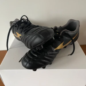 Svarta fotbollsskor från Mizuno PRO FG/AG - Snygga svarta fotbollsskor från Mizuno med guldiga detaljer och snörning. De är knappt använda bara fåtal gånger. Ser helt oanvända ut.  Storlek 42,5. Skorna är även FG/AG som står för firm ground och artificiall grass så skorna går och användas på alla olika sorters fotbollsplaner. De är Pro och kostade ca 1300 kr nya. Vill snabbt få sålt därför det låga priset. 