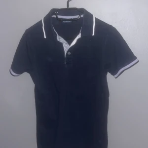 Svart pikétröja med vita detaljer - Snygg svart pikétröja  med vita detaljer på krage och ärmslut. Perfekt för en stilren look. Tröjan har en klassisk pikékrage och knappar framtill.