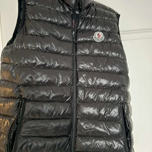 Svart dunväst från Moncler - Snygg svart dunväst från Moncler med dragkedja och logotyp på bröstet. Perfekt för kyligare dagar och ger en stilren look. Västen har en glansig yta och praktiska fickor med dragkedja. Äkta med fungerade QR kod. Som ny💯👍